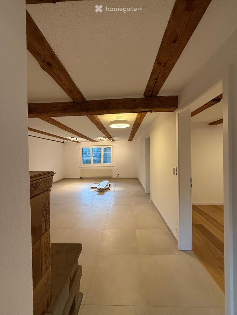Empty room with exposed wooden beams, tiled floor, and a window with a radiator below it. The room has a simple, clean design with white walls and a recessed ceiling light.