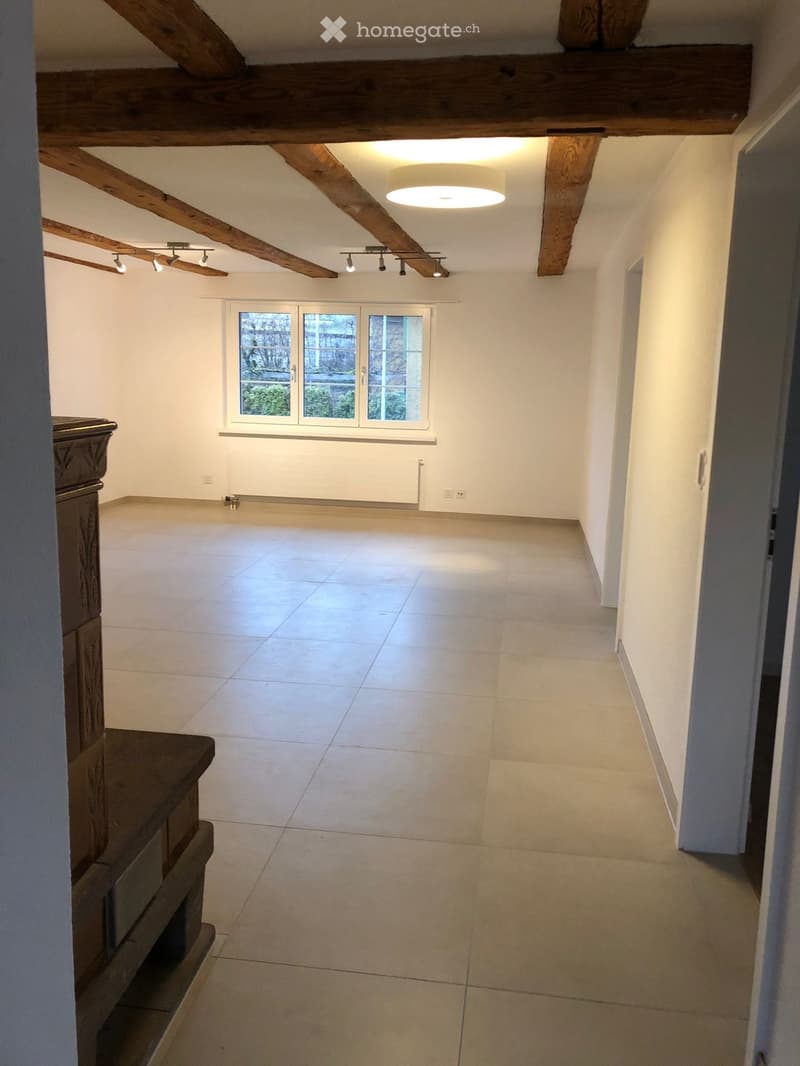Empty room with exposed wooden beams, tiled floor, large window, and modern lighting fixtures.