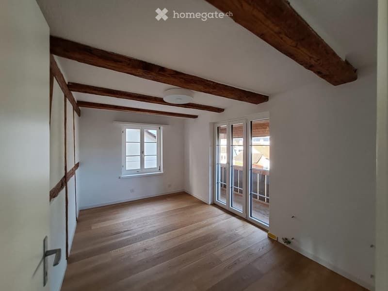 Empty room with wooden floor, exposed wooden beams on the ceiling, white walls, and a large window with white frames. A sliding glass door leads to a balcony or terrace area.