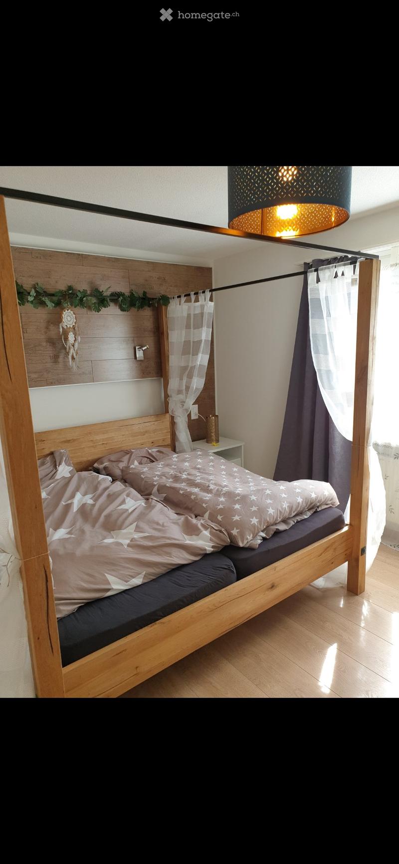 A cozy bedroom with a wooden bed frame, a nightstand, and a hanging lamp. The bed is made with patterned bedding, and there is a dreamcatcher decoration on the wall.
