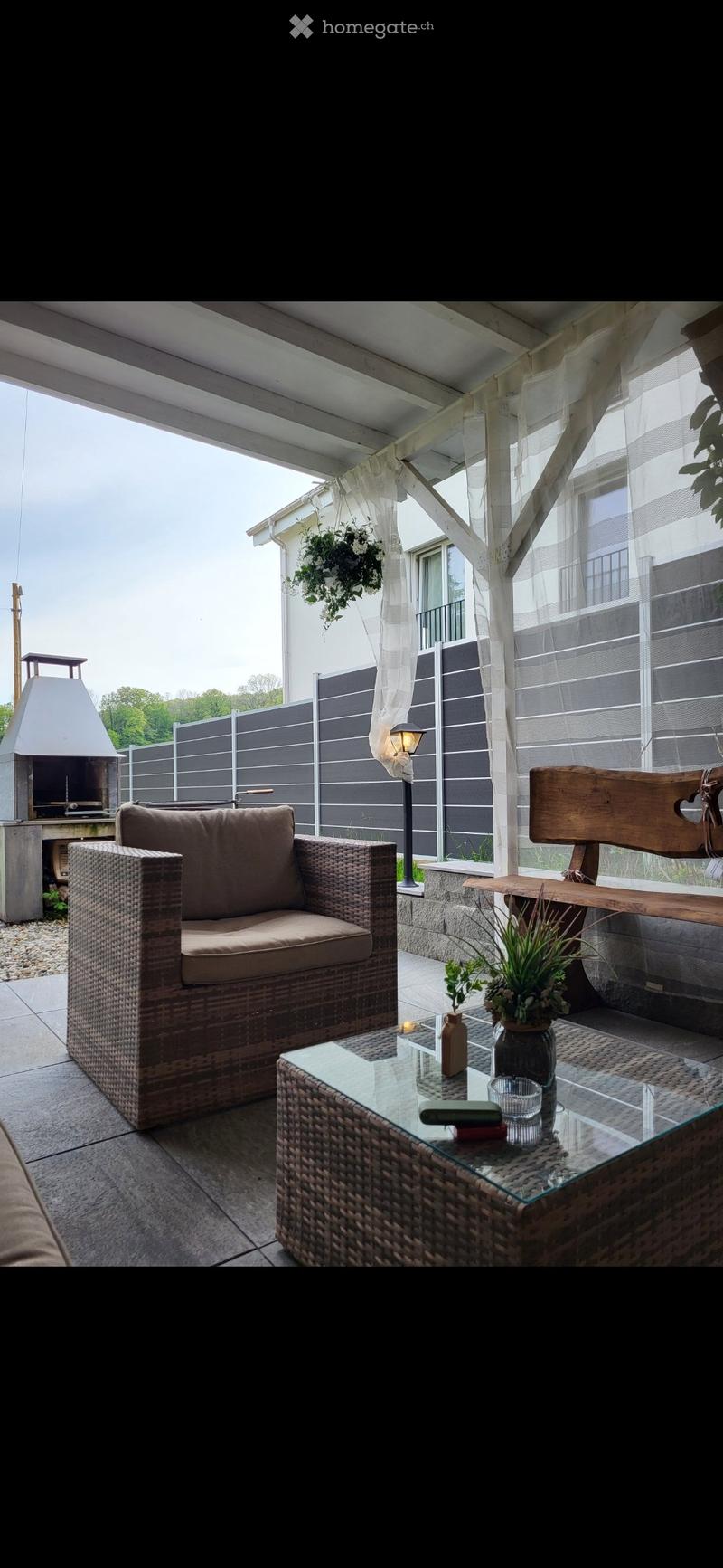 A cozy outdoor patio with a wicker sofa, glass coffee table, wooden bench, and hanging plants.