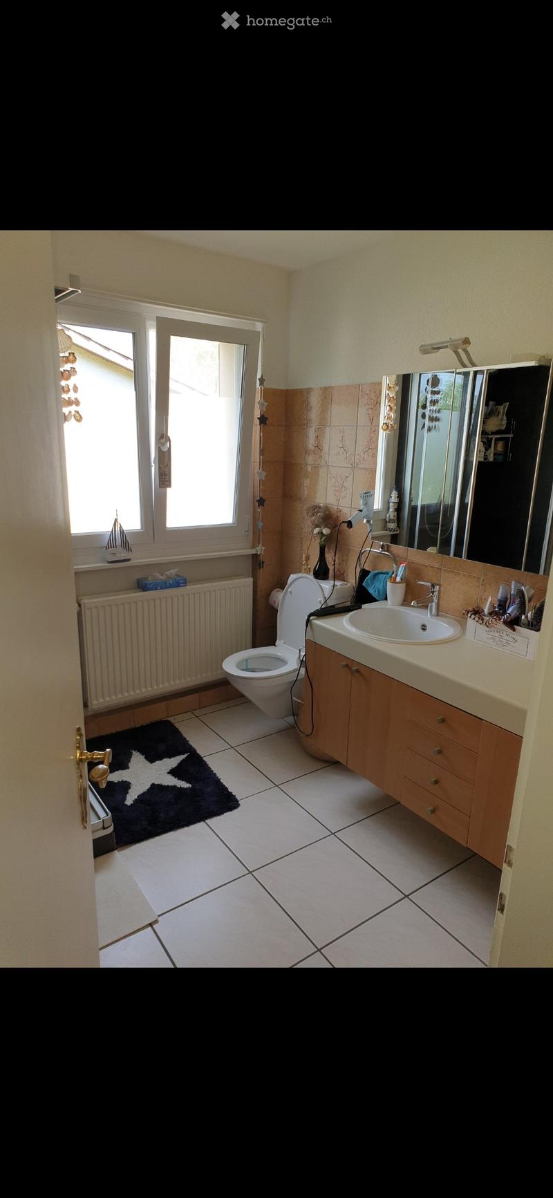 Bathroom with white tiled floor, beige walls, sink with wooden cabinet, toilet, mirror, and a small window with a decorative sailboat on the sill.