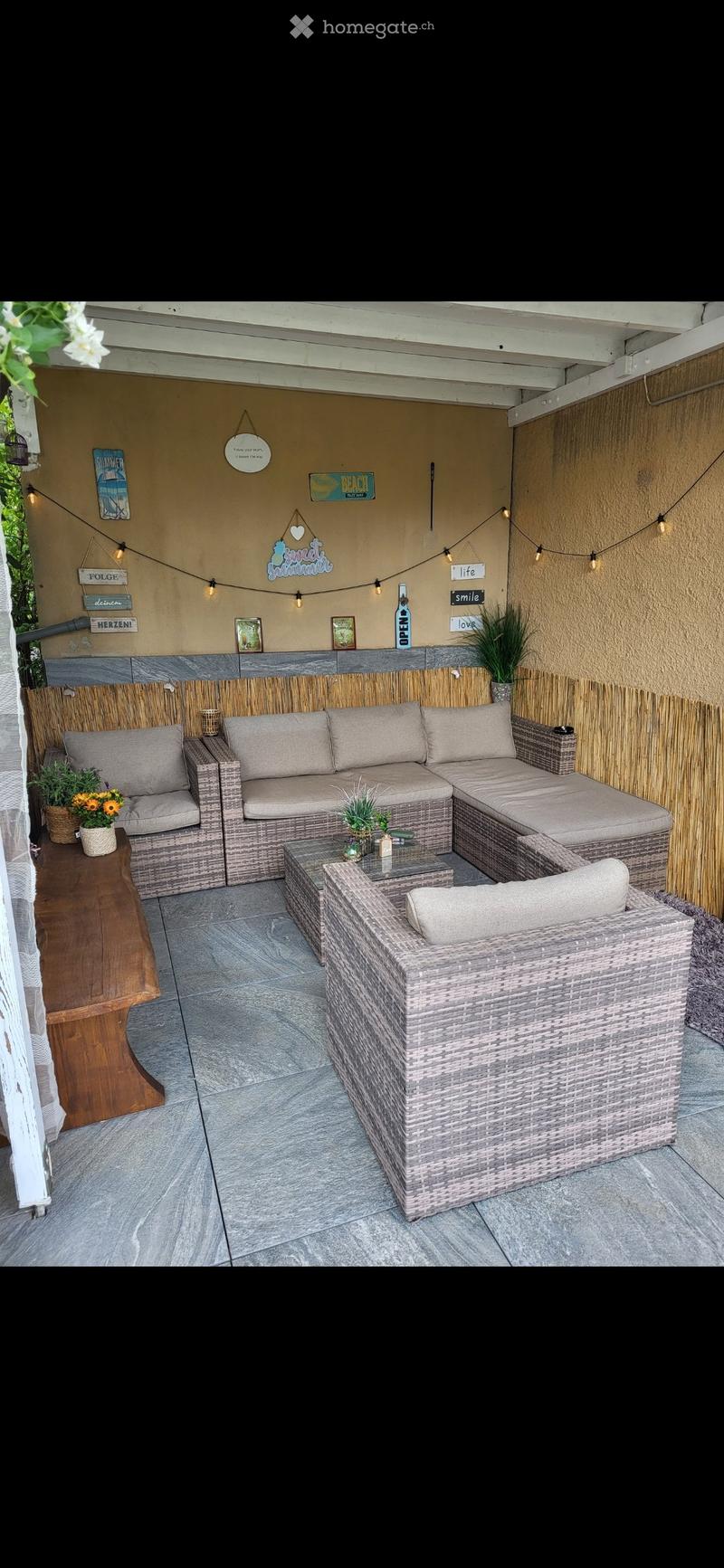 A cozy outdoor patio with a wicker sofa set, a wooden side table, and a small coffee table, decorated with string lights and potted plants.