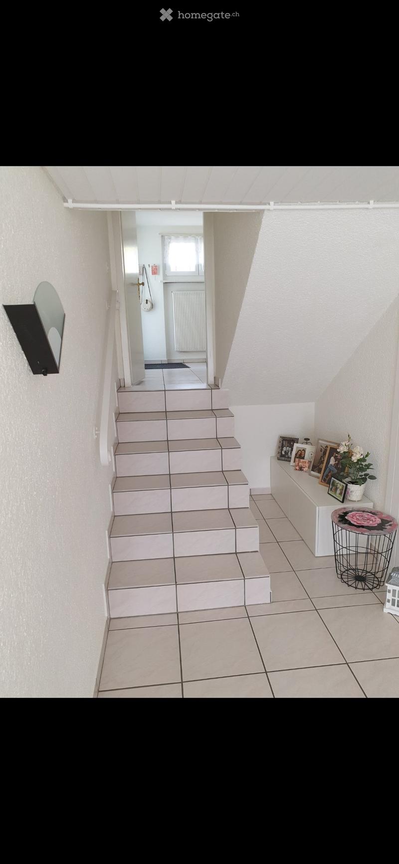 Hallway with tiled steps leading to another room, small table with picture frames and a flower pot, and a round stool nearby.