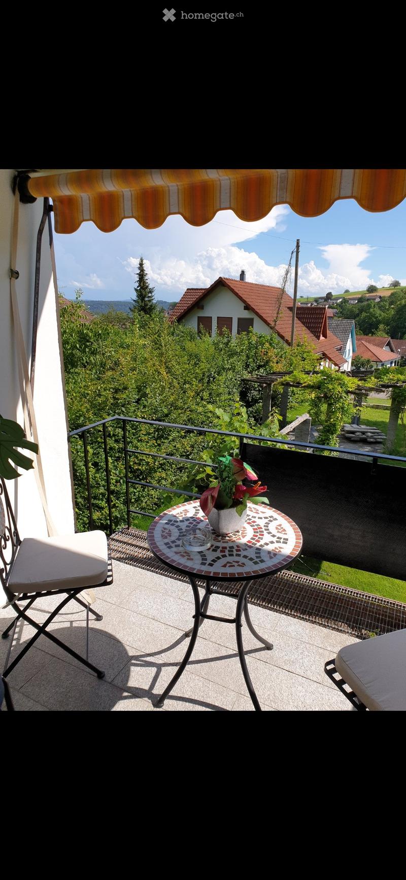 Balcony with a round mosaic table, a white chair, and a flower pot, overlooking a green landscape with houses and trees.