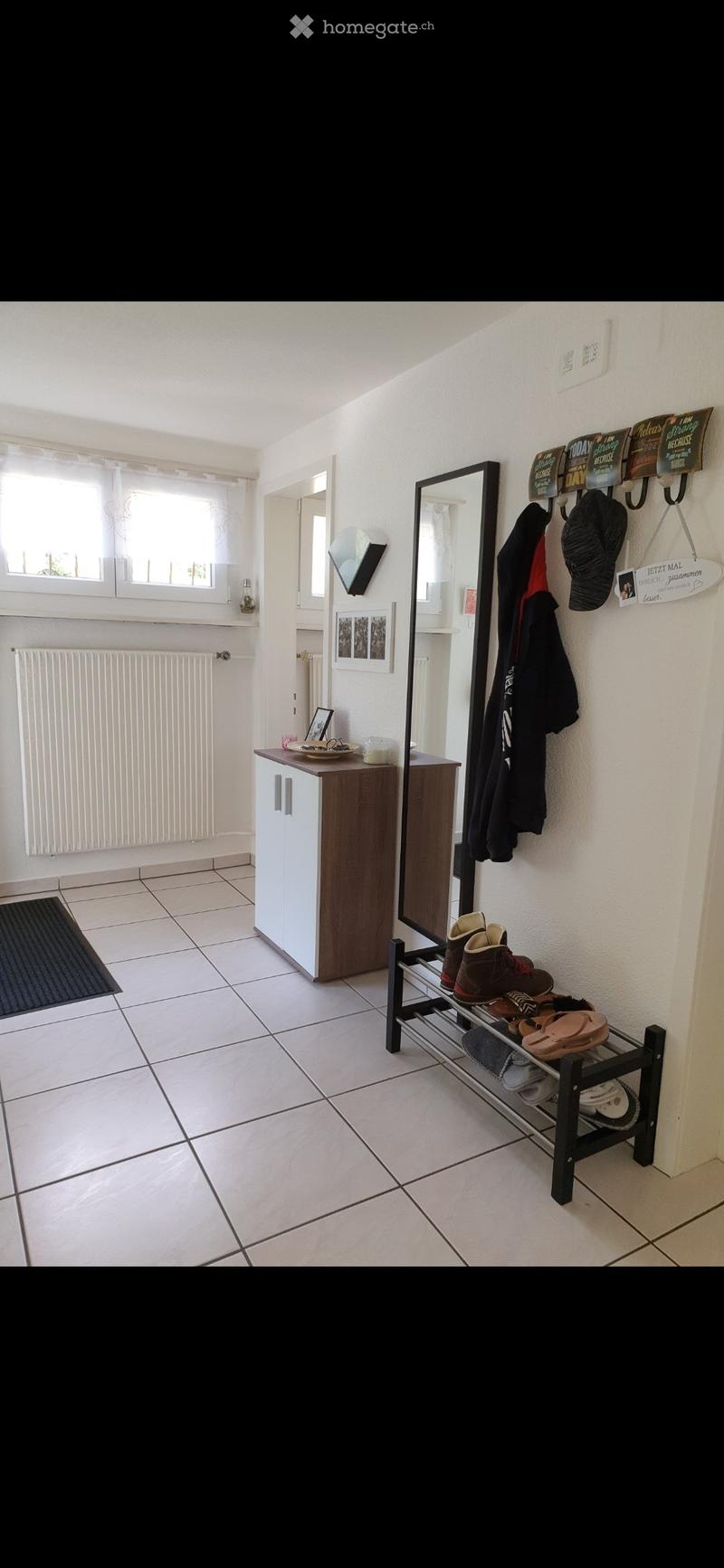 Bright hallway with tiled floor, white walls, coat rack, shoe rack, small cabinet, and mirror. Natural light from windows on the left side.