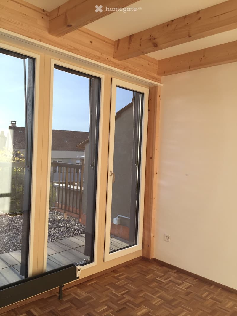 Empty room with wooden herringbone flooring, white walls, and large glass doors leading to a balcony area with a wooden railing and tiled floor.