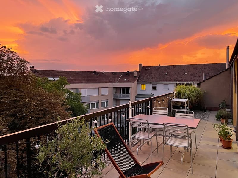 Balcony with a table, chairs, potted plants, and string lights, overlooking a building with a sunset sky.