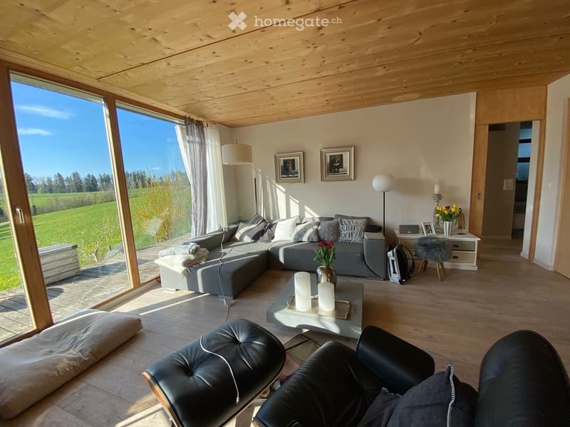 Modern living room with large windows overlooking a green field, grey sectional sofa, leather chairs, coffee table, floor lamp, and decorative items.