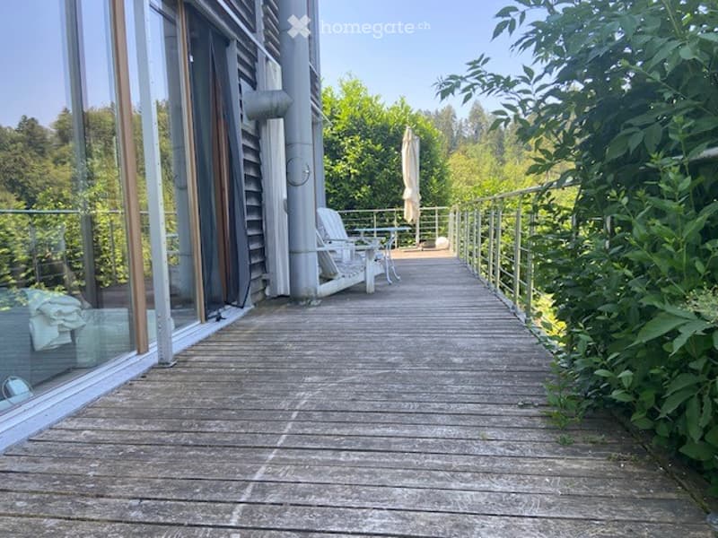 Wooden deck with glass railing, leading to a white lounge chair and small table, surrounded by greenery and trees.