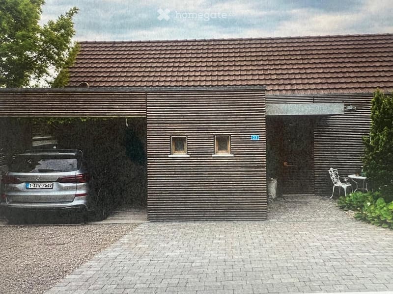 Modern two-story house with a dark wooden facade, tiled roof, and a covered garage. A silver car is parked inside the garage, and there is a small outdoor seating area with a white metal chair and table near the entrance.
