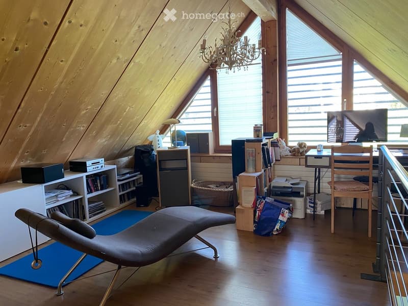 Attic room with wooden slanted ceilings, large windows, a lounge chair, a desk with a screen, shelves with books and electronics, and a chandelier.