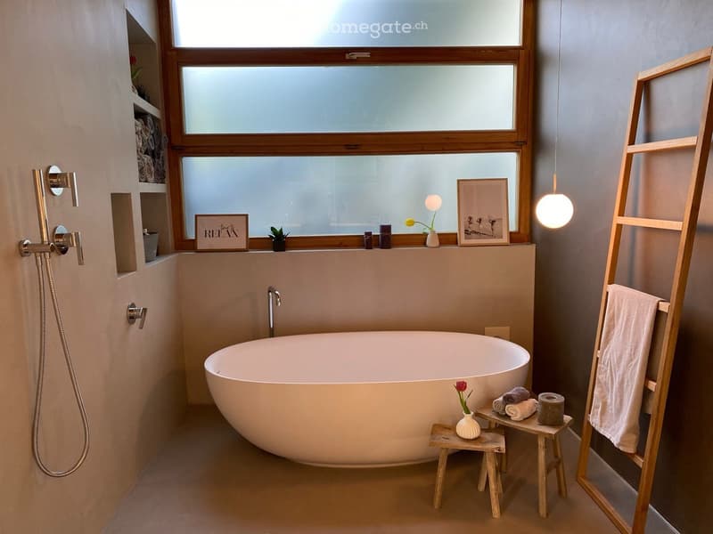 Modern bathroom with a freestanding bathtub, wooden stool, small vase with flower, towel rack, and decorative items on the windowsill.