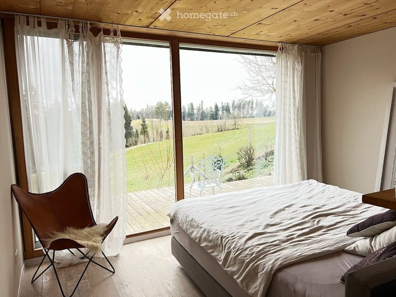 Bright bedroom with large windows, wooden floor, white bed with pillows and blanket, brown chair with fur throw, and a view of a green field and trees.