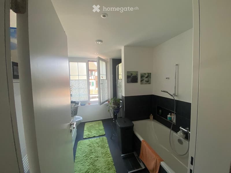 Modern bathroom with a bathtub, toilet, shower, and green rugs. Features a window with a view of the outside, a small plant, and decorative pictures on the wall.