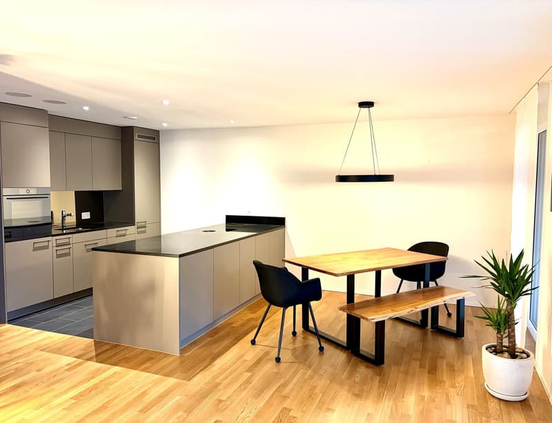 Modern kitchen with a central island, wooden table, black chairs, bench seating, and a potted plant. Features grey cabinets, a black countertop, and a pendant light fixture.