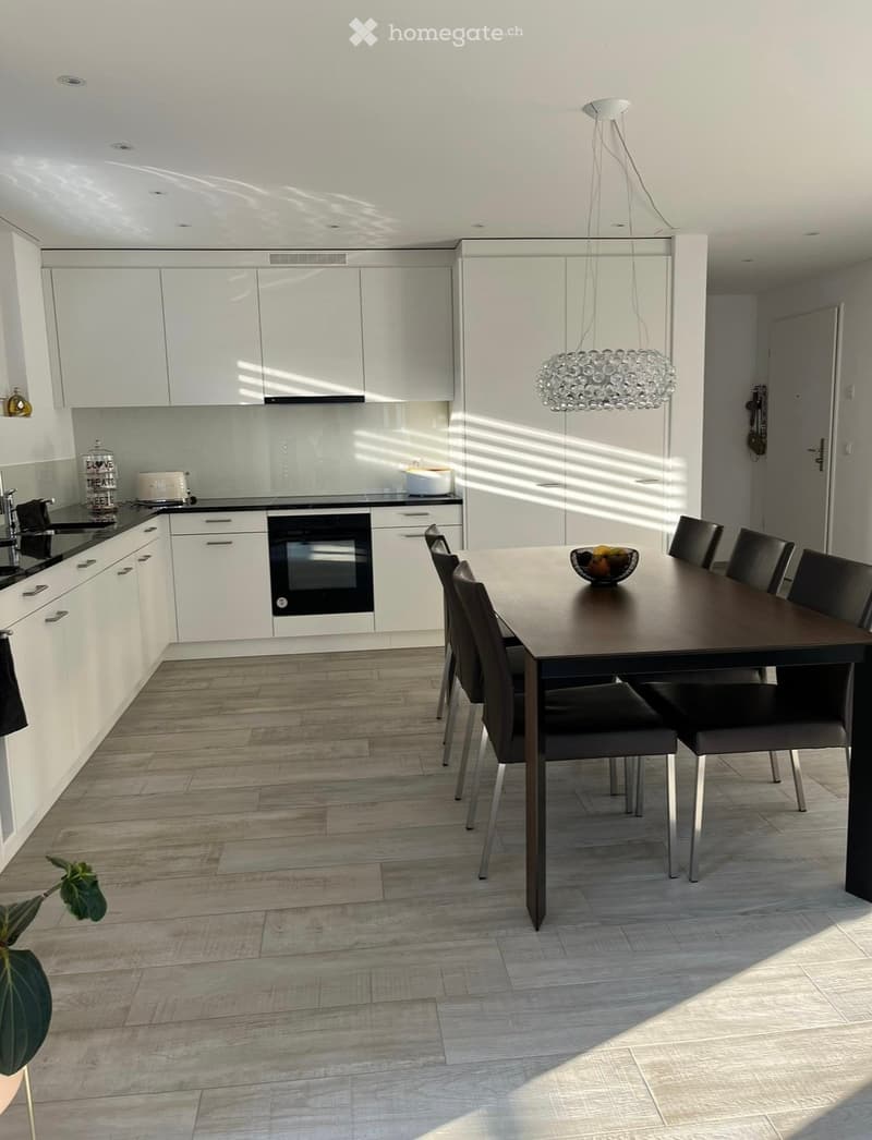 Modern kitchen and dining area with white cabinets, dark countertops, a wooden dining table with six chairs, and a hanging crystal chandelier.