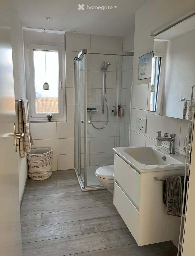 Modern bathroom with a glass-enclosed shower, white sink with chrome faucet, toilet, and a wicker basket near the window. The room features light-colored tiles and wooden flooring.