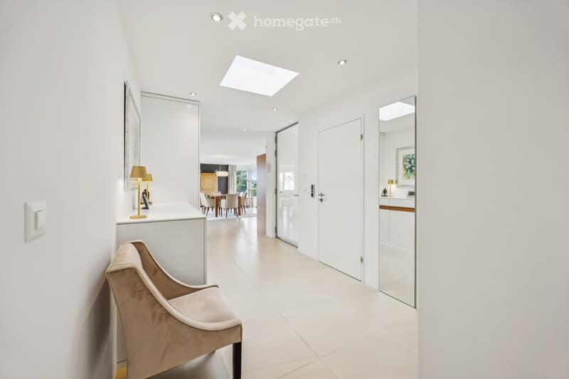 Corridor / Hallway. Bright, modern hallway with white walls, recessed lighting, and a skylight. Features a small seating area with a chair and a console table with lamps, leading to a dining area visible in the background.