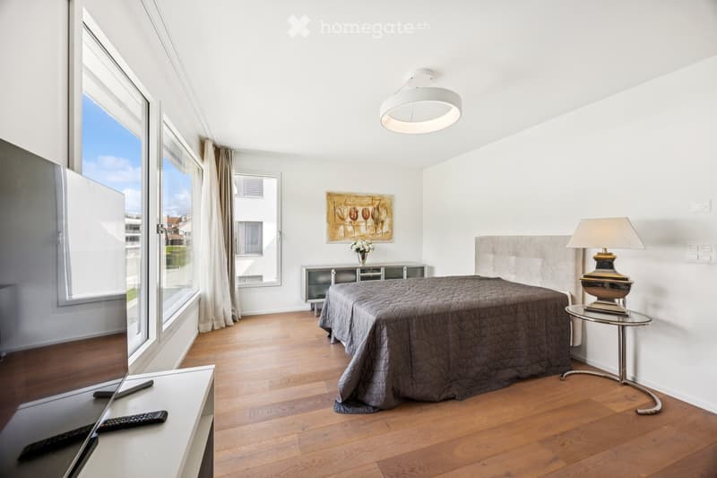 Bedroom. Modern bedroom with wooden flooring, large windows, a bed with a grey duvet, a nightstand with a lamp, a small table, and a painting on the wall.