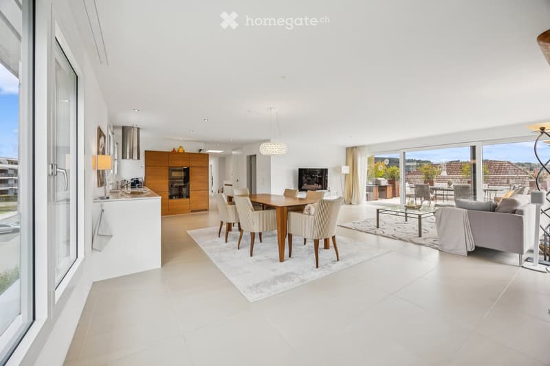 Furnished Room. Modern open-plan living and dining area with white walls, large windows, and a view of the landscape. Features include a grey sofa, coffee table, dining table with chairs, kitchen area with cabinets and an oven, and a television. The space
