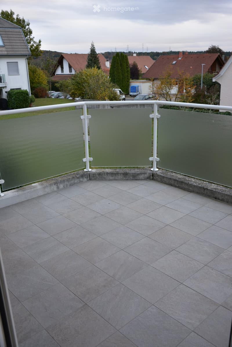Balcony with grey tiled floor, white metal railing, and glass panels, overlooking a residential area with houses and trees.