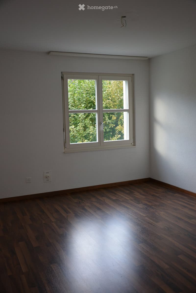 Empty room with wooden flooring and a large window letting in natural light. The walls are plain and there are no furnishings or decorations visible.