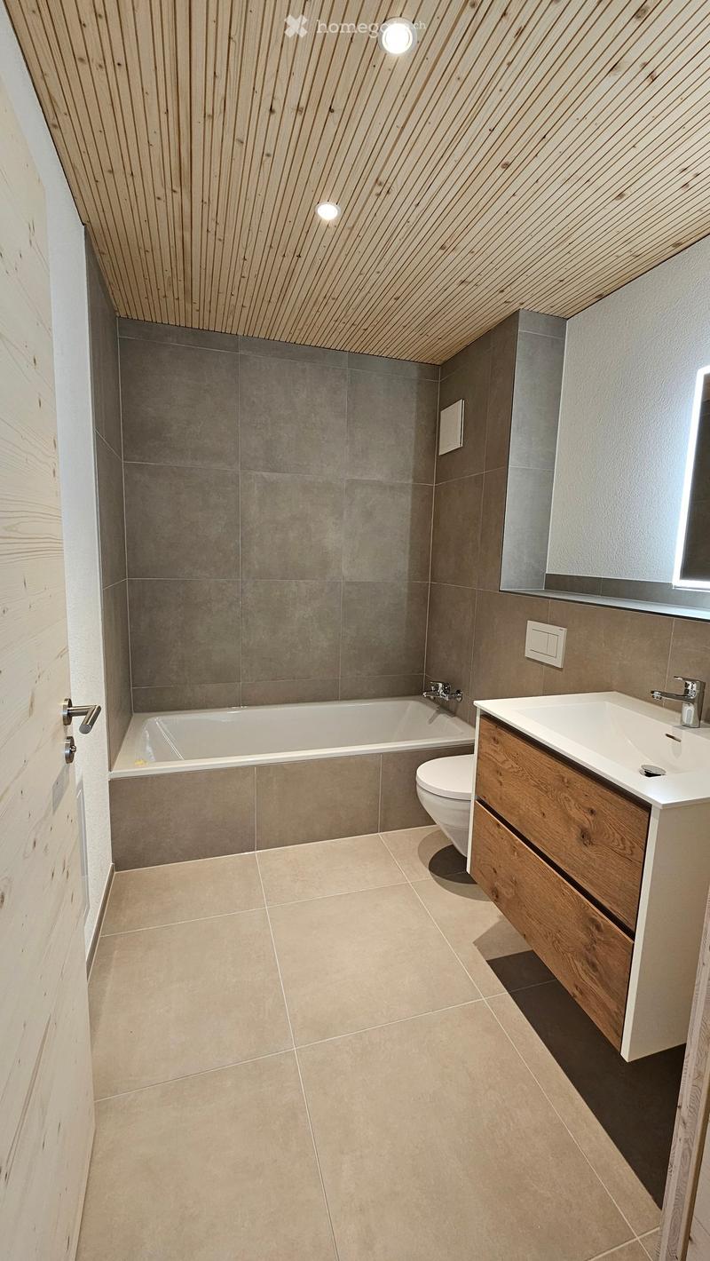 Modern bathroom with a bathtub, toilet, sink, and wooden cabinet. Features light-colored tiles and a sleek wooden ceiling.