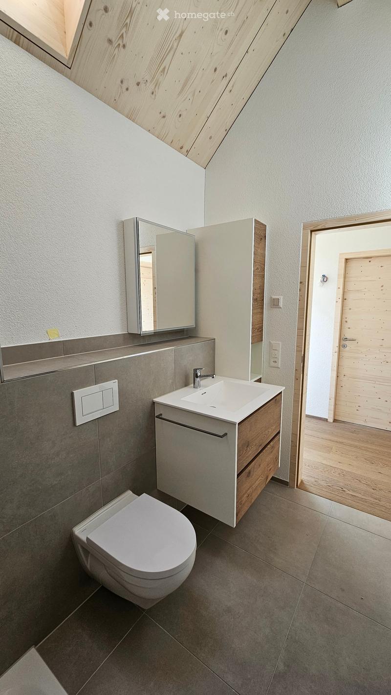 Modern bathroom with a white sink, chrome faucet, mirrored cabinet, and a toilet. The walls are finished in a textured grey tile, and the floor is made of large, light grey tiles.