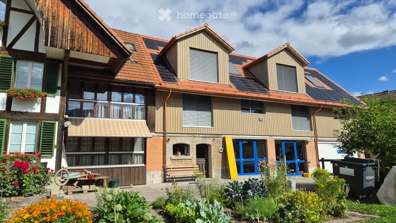 A multi-story residential building with a mix of modern and traditional architectural elements, featuring solar panels on the roof, large windows, and a well-maintained garden area with flowers and plants.