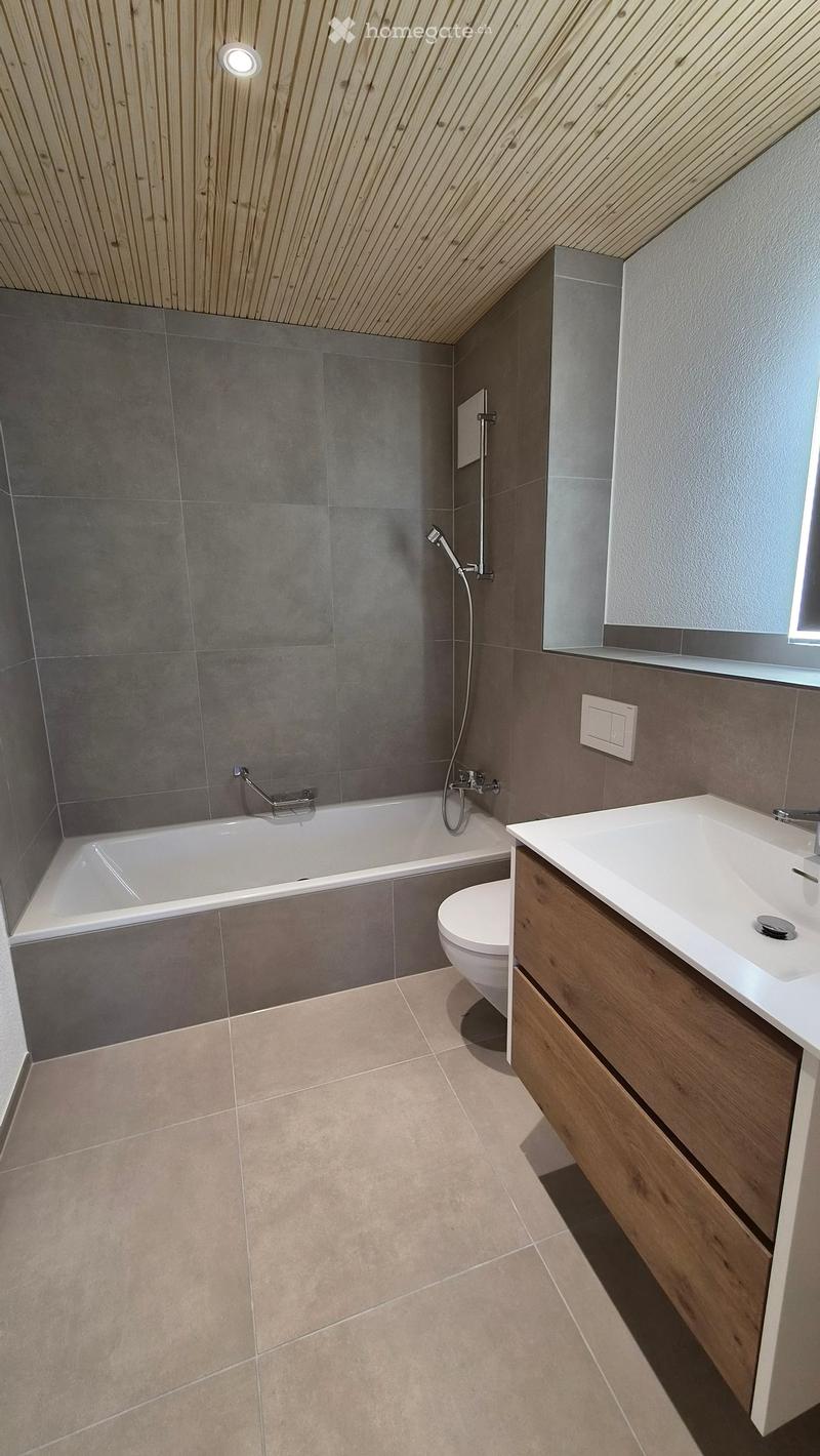 Modern bathroom with a bathtub, toilet, sink, and mirror. Grey tiled walls and light wooden cabinetry.
