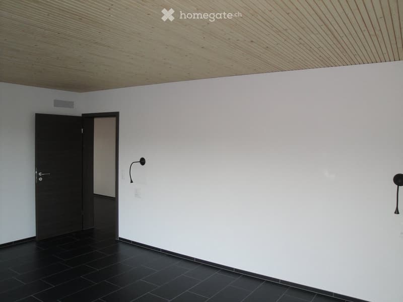 Empty room with white walls, black tiled floor, and a wooden slatted ceiling. Features a black door, wall-mounted light fixture, and a small vent on the wall.