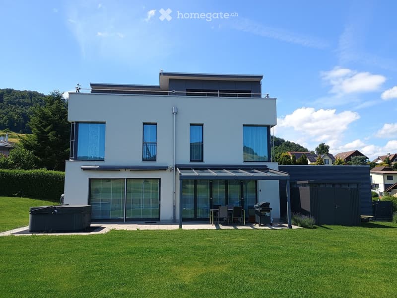 Modern two-story house with light blue exterior, large windows, covered patio area, outdoor seating, grill, and hot tub in a well-maintained lawn.