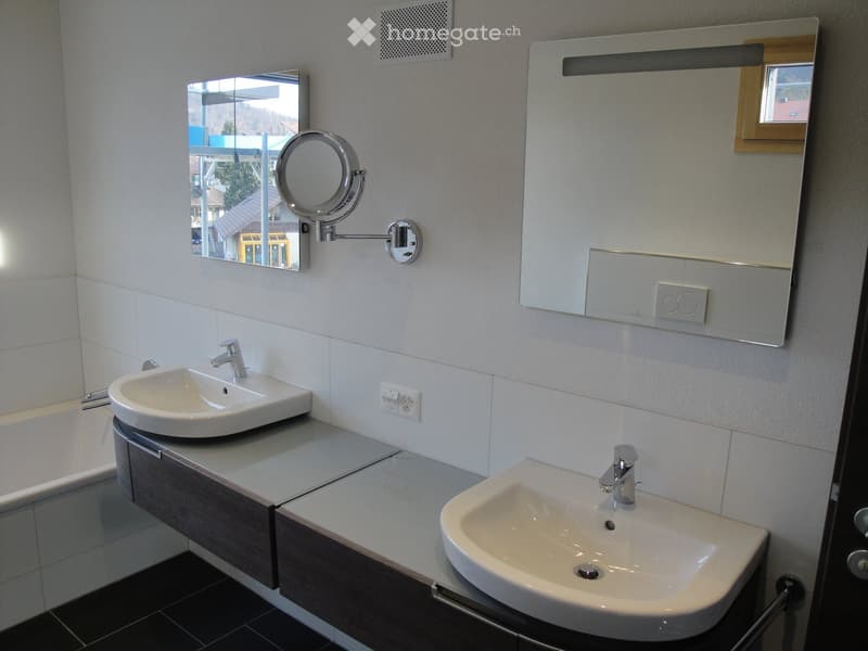 Modern bathroom with two sinks, a vanity cabinet, and a large mirror with built-in lighting. The walls are tiled in white, and the floor is dark.