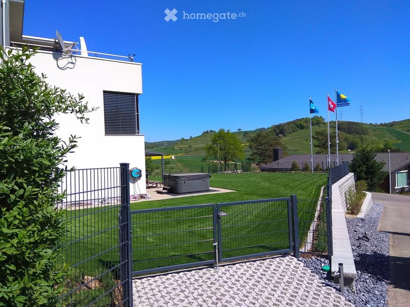 A modern house with a well-maintained lawn, a hot tub, and a metal gate. The background features rolling hills and flags on poles.