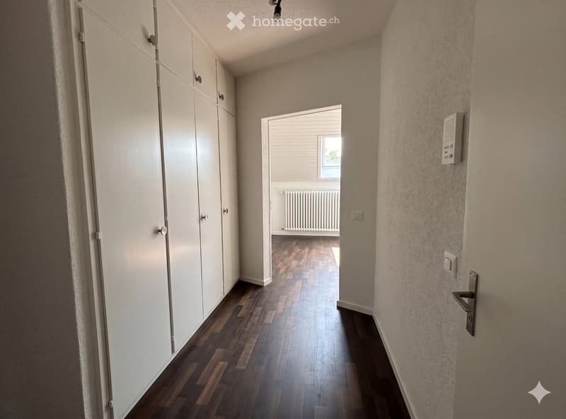 A narrow hallway with wooden flooring, white walls, and built-in white cabinets on one side. A door at the end leads to another room with a radiator and a window.