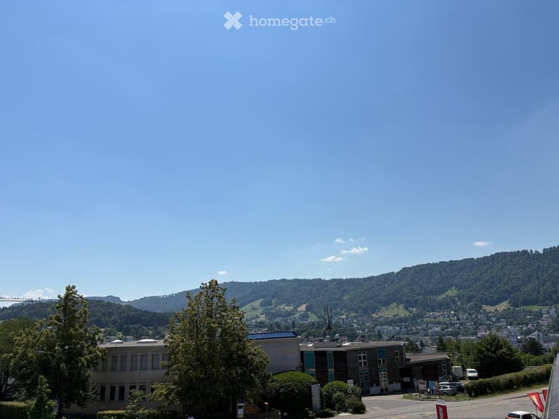 Modern building with multiple windows and a flat roof, surrounded by trees and situated in a scenic valley with hills in the background.