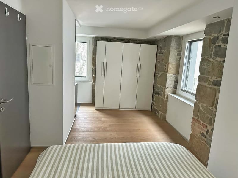 Bedroom with a striped bedspread, a large white wardrobe, wooden flooring, and exposed stone walls. The room has large windows and a door leading to another area.
