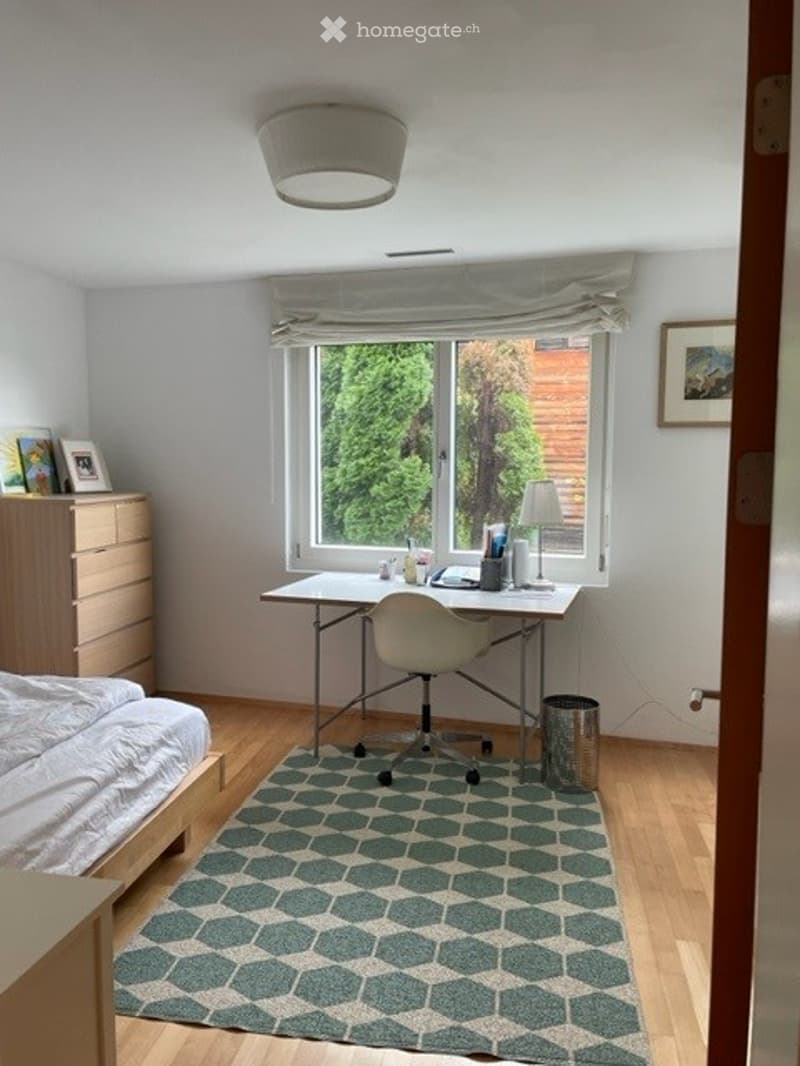 Cozy bedroom with a bed, desk, chair, and a patterned rug. The room features a large window with a view of greenery, a ceiling light, and a small lamp on the desk.