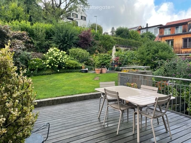 A wooden deck with a dining table and chairs, surrounded by a well-maintained garden with various plants and trees. The background shows a residential area with houses and greenery.