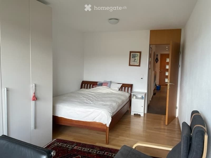 Cozy bedroom with wooden flooring, a neatly made bed, a nightstand, a chair, and a picture frame on the wall.