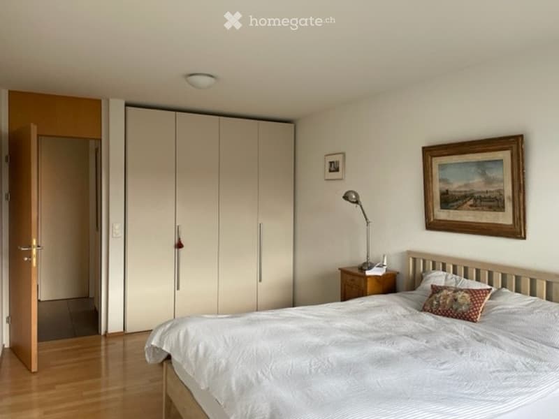 A well-lit bedroom with a large bed, nightstand, lamp, wardrobe, and framed artwork on the wall.