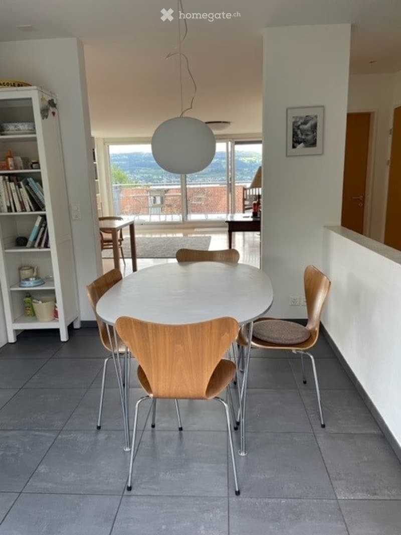 Modern dining area with a white round table, six wooden chairs, a white bookshelf, and a large pendant lamp hanging from the ceiling. The room has large windows offering a scenic view.