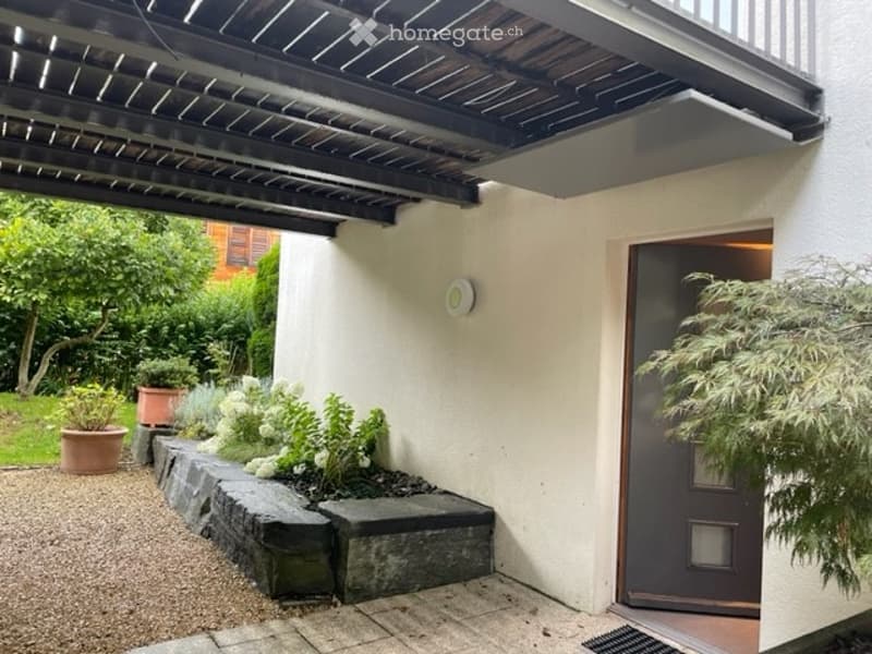 A small, well-maintained garden area adjacent to a house entrance, featuring potted plants, a low stone wall, and a covered walkway.