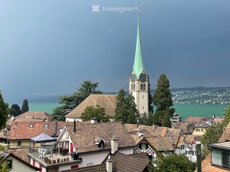 A picturesque town view with a prominent church tower featuring a green spire and clock faces, surrounded by traditional houses with tiled roofs and lush greenery, overlooking a serene lake and distant mountains.
