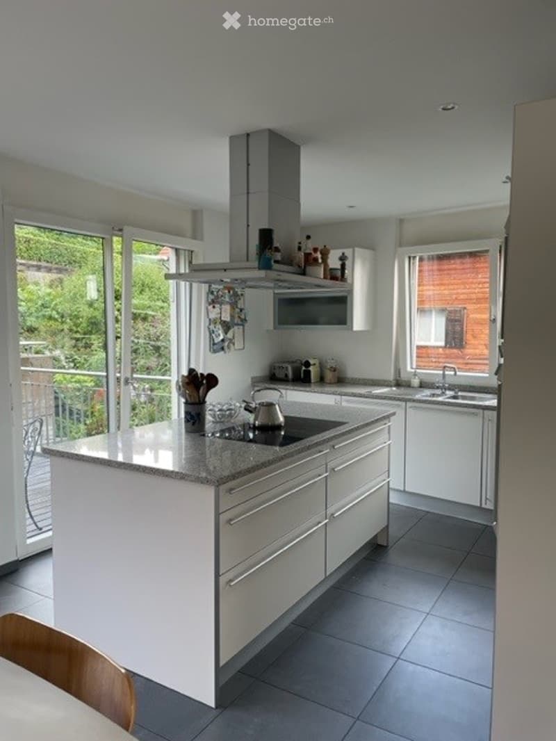 Modern kitchen with white cabinets, island, induction stove, and large windows offering a view of greenery.