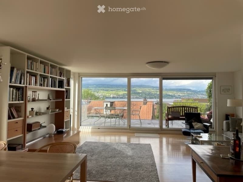 Bright living room with large windows offering a scenic view, furnished with a sofa, coffee table, bookshelf, and a dining area.