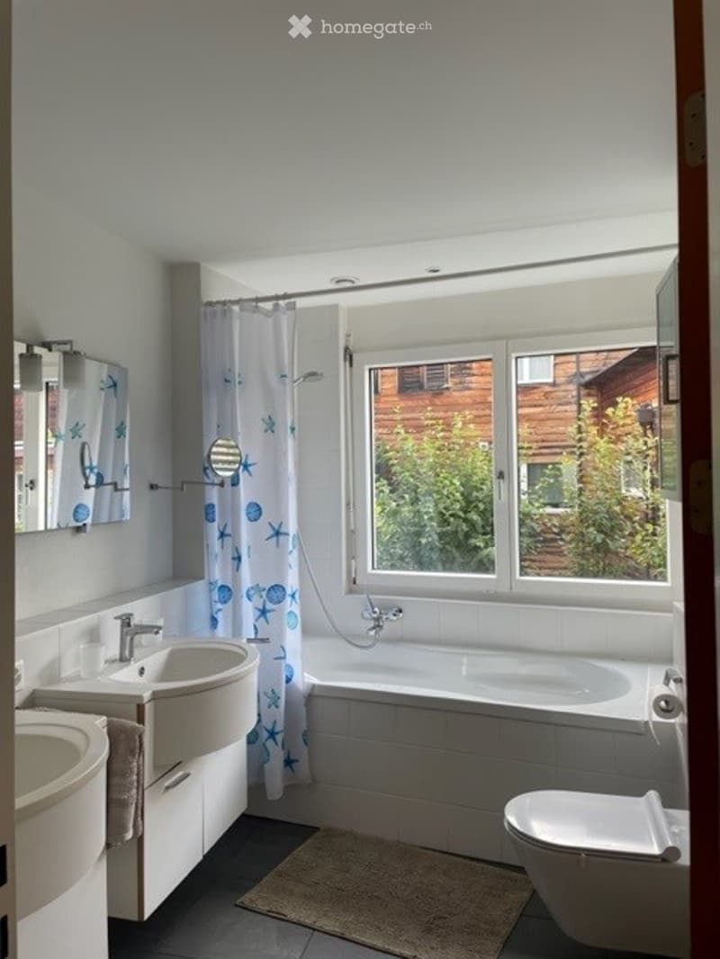 Modern bathroom with white fixtures, a bathtub, two sinks, a toilet, and a shower curtain with a blue sea theme. A large window provides natural light.