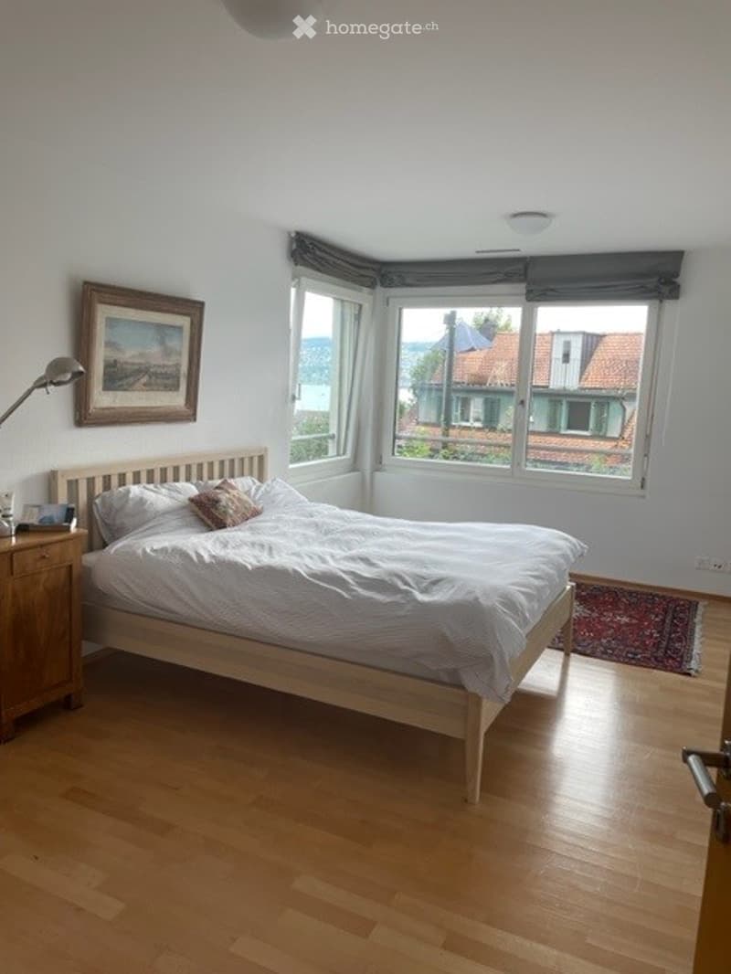 Bright bedroom with wooden flooring, large windows, a neatly made bed with pillows and a blanket, a nightstand with a lamp, and a framed picture on the wall.