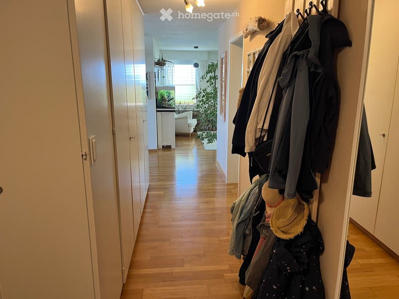 A well-lit hallway with wooden flooring, white walls, and several doors leading to other rooms. A coat rack with various jackets and hats is visible on the right side.